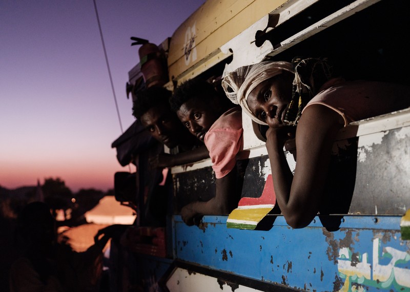 A group of people are on a bus, which is backlit by an ominous purple blue and red sky. The expressions on the travellers faces show distress, anxiety and loss.