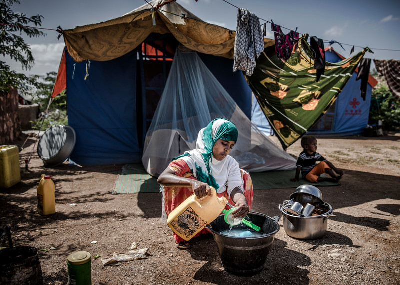 a young woman washes dishes in a bucket outside a blue tent that is draped in colourful fabrics. There is a laundry line hung out the front and a little boy sitting on the ground staring into the distance