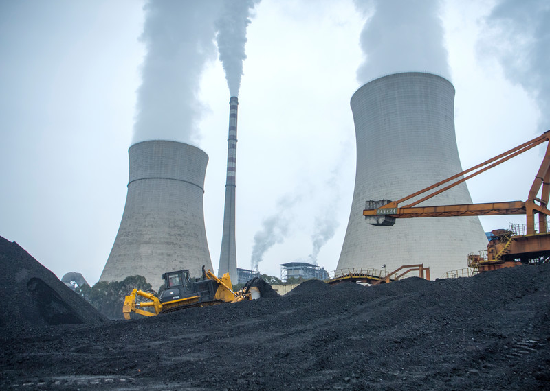 Two enormous pillars at the Giangyou Power station in China. In front of them, yellow bulldozers and cranes crawl across dark and sandy hills of coal.  