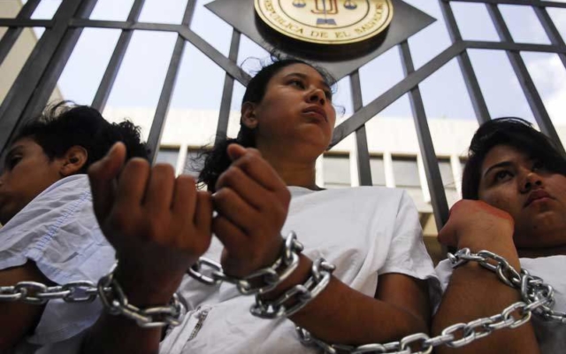 Three women in white shirts stand in front of a gate, their arms chained together. Their glances focus behind the camera in defiance. 