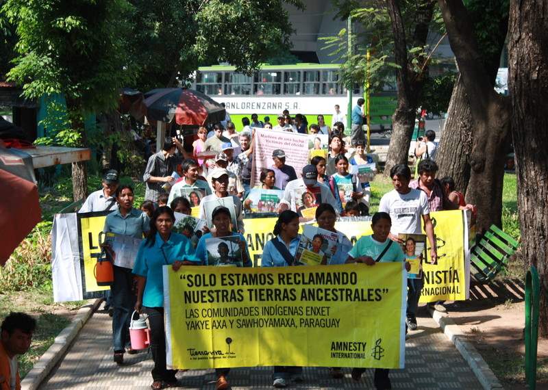 a group of protestors parading down a pedestrian walkway. They are holding large amnesty banners with calls to support the Yakye Aza and Sawhoyamaxa Indigenous communities in Paraguay. 